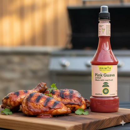Bottle of Primor Foods Pink Guava syrup next to grilled chicken on a wooden cutting board with a blurred grill in the background.